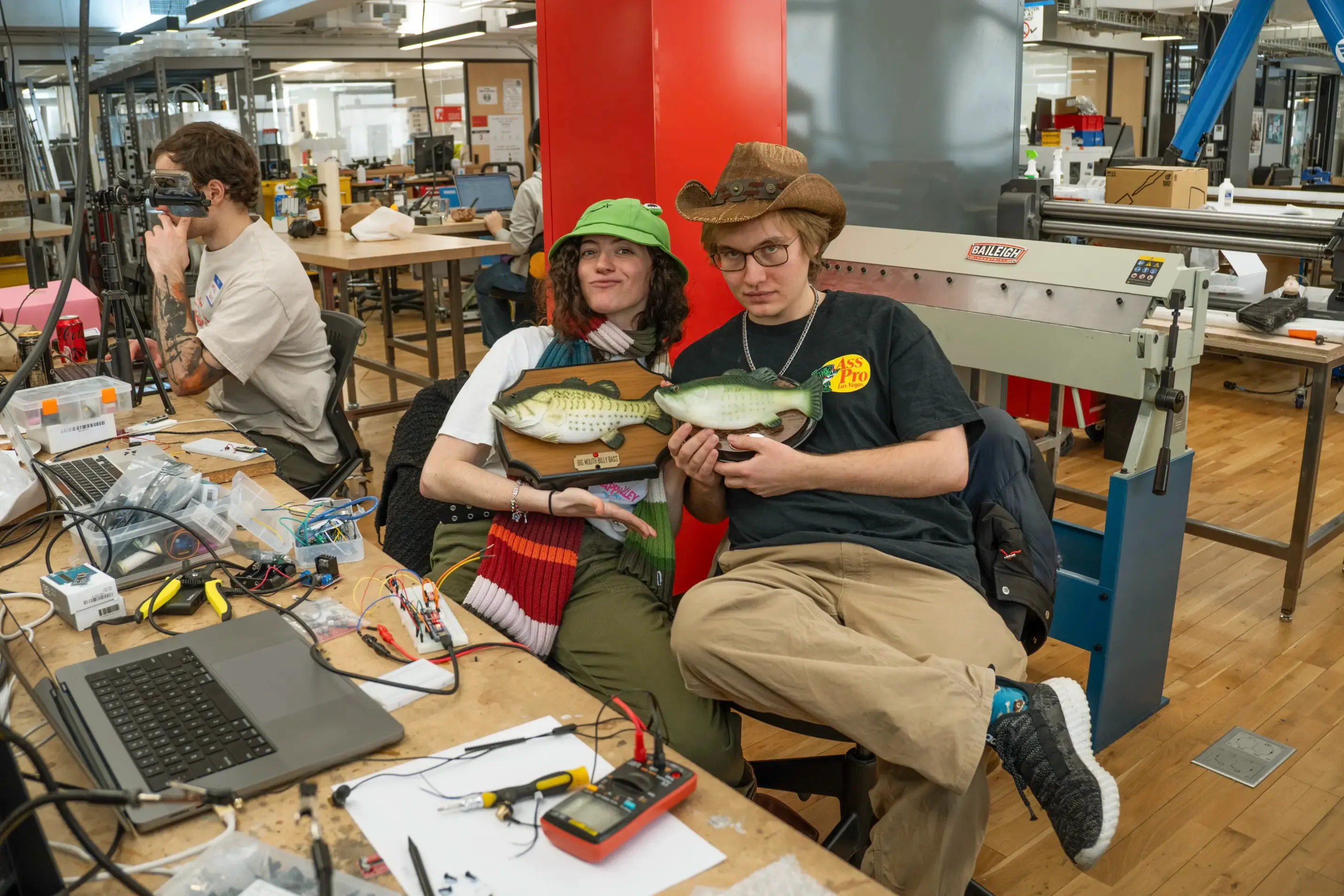 Portrait of two hackers holding plastic fish, one wearing a frog hat and the other a cowboy hat, in front of soldering equipment and laptops in the workshop