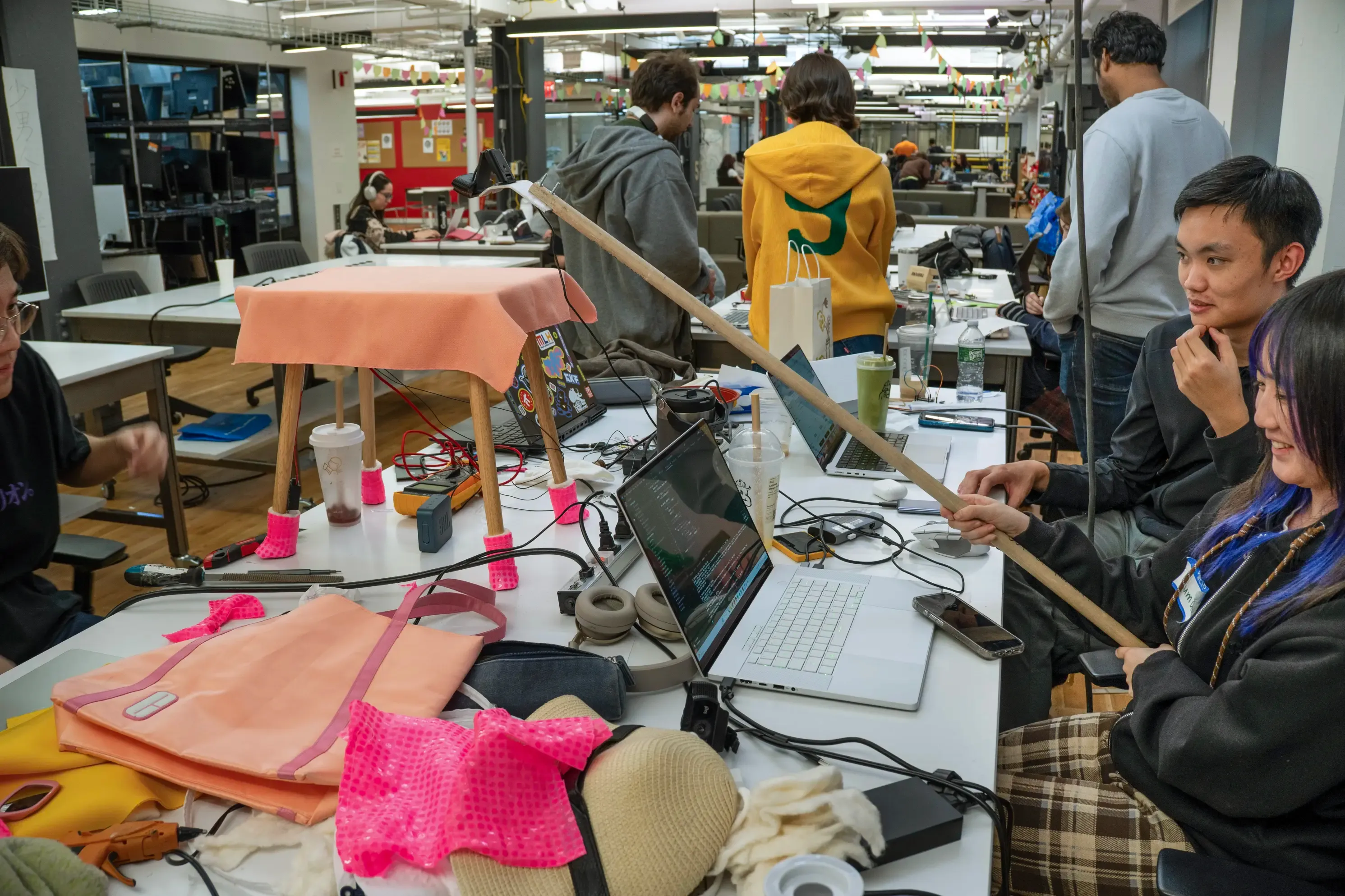 3 students hold a camera on a dowel over a table wearing hot pink booties, surrounded by a laptop and fabric and electronics