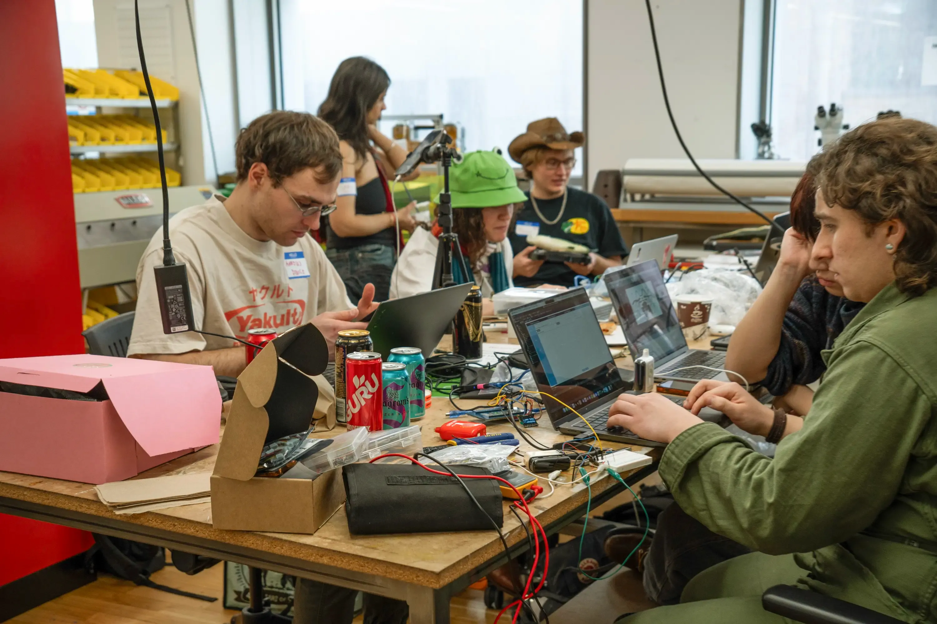 Hackers in the fabrication studio, with the nearest hacker wearing a green jumpsuit surrounded by vapes hooked up to multimeters and an Arduino